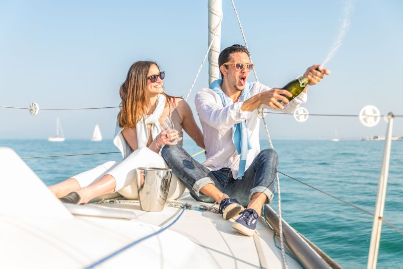 Couple celebrating anniversary with champagne on a boat - Attractive caucasian man uncorking champagne and having party with her girlfriend on vacation - Two young tourists having fun on a boat tour in the summertime